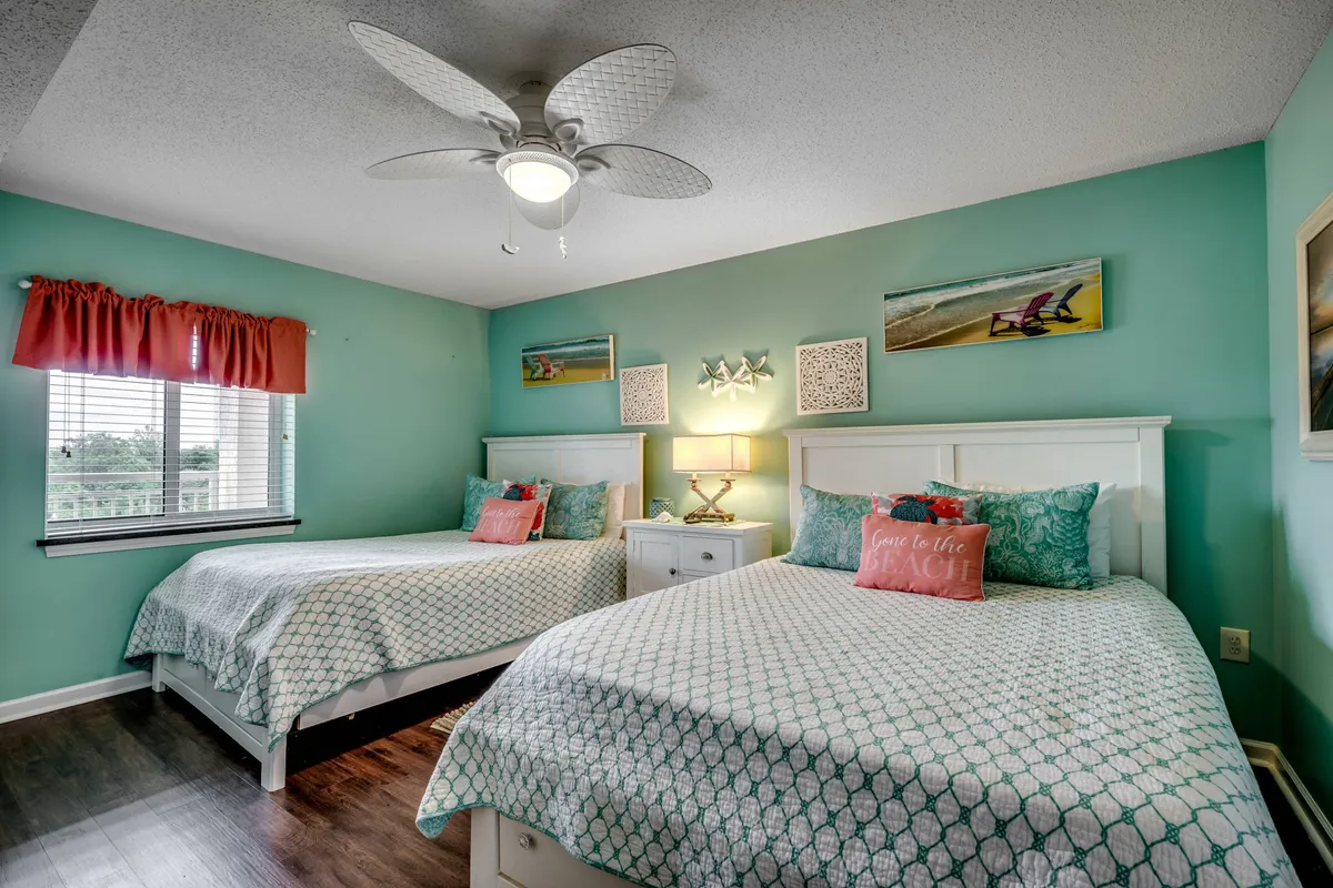 A welcoming guest suite furnished with two comfortable beds topped with decorative pillows and soft linens. A window with curtains lets in gentle light, and the bedside area offers a cozy spot for reading or sharing stories, reflecting a family-friendly ambiance.