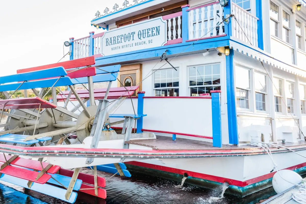An exterior shot of the Barefoot Queen Riverboat, showing its paddlewheel, colorful decor, and signage. The boat is docked and ready for guests, with festive lighting suggesting an atmosphere of lively entertainment on the water.