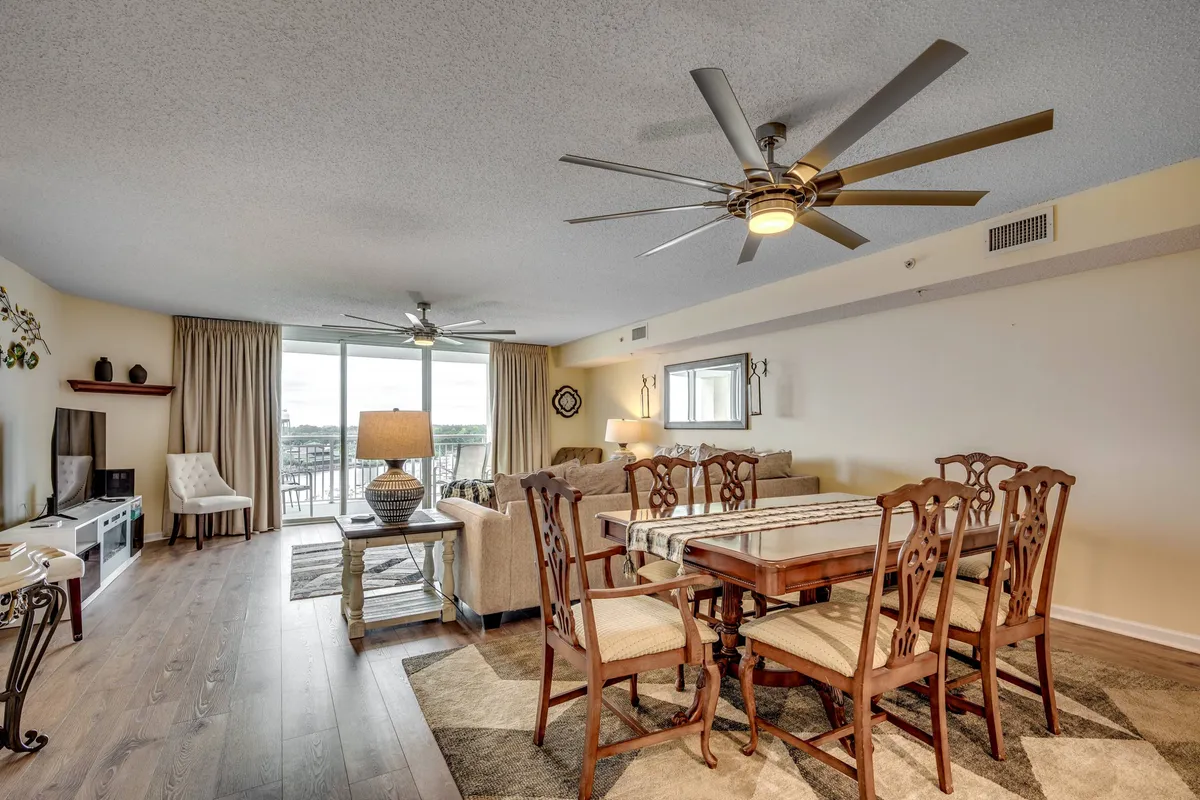 A spacious open-concept interior featuring a dining table set for a meal, comfortable living room seating, ceiling fans above, and large windows that offer beautiful views. The sunlight streaming in creates a welcoming and cozy atmosphere ideal for gatherings.