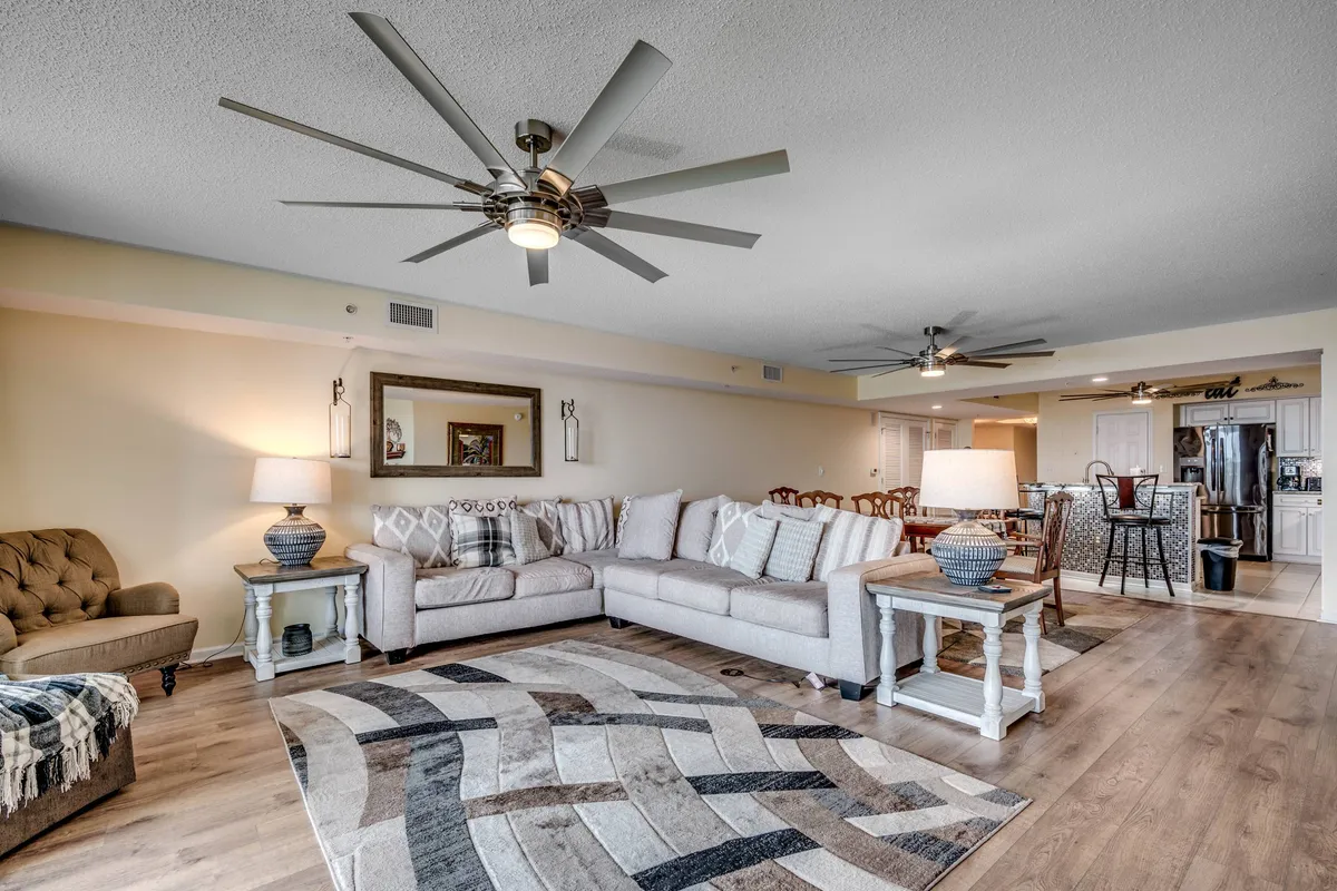 The image showcases a spacious and inviting living area within the condo, featuring a large, comfortable sectional sofa arranged around a plush, stylish area rug. Decorative lamps illuminate the room with a warm glow, while a framed mirror adds depth and elegance to the space. The setting is designed for relaxation, socializing, and quality time with family or friends.