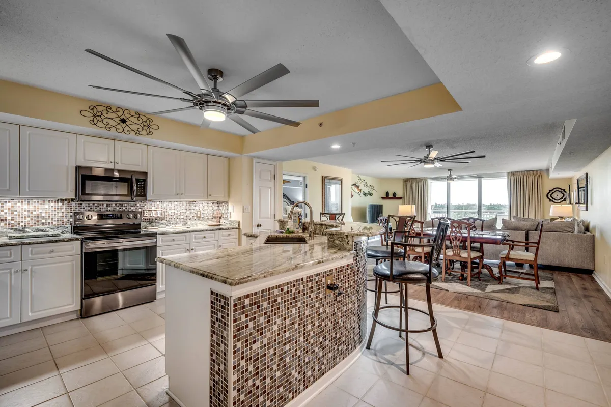 A stylish kitchen island with stainless steel appliances opens seamlessly into an inviting living area and adjacent dining space. Natural light fills the room, and the layout encourages easy movement between cooking, dining, and lounging.