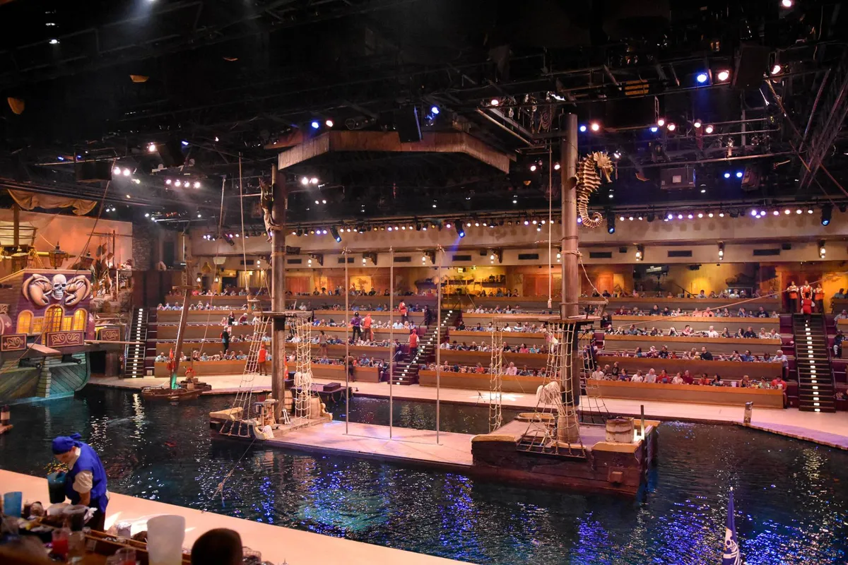 This interior action shot captures the Pirate's Voyage Dinner & Show. The scene features a large stage area complete with a pirate ship floating on water, dramatic lighting, and an audience seated and engaged with the high-energy performance.