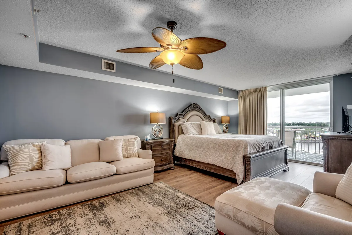 A tranquil primary suite featuring a plush king-sized bed with soft linens, a comfortable sofa for lounging, ceiling fan overhead, and balcony doors that allow natural light to flood the room. The space exudes a relaxing atmosphere, perfect for unwinding after a busy day.